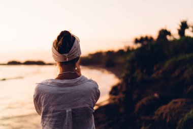 Okyanusta günbatımının tadını çıkarırken, kumlu tepede çörekler içinde saçları olan tanınmayan kadın kaşifin arka görüntüsü.