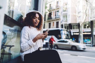 Düşünceli Afro-Amerikalı kadın, günlük giysiler ve şapkayla ahşap bankta oturmuş, akıllı telefonları karıştırıyor ve caddede dinleniyor ve kameraya bakıyor.