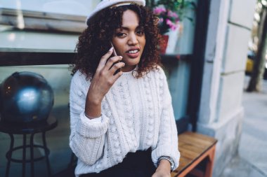 Sıradan giysiler ve kol saati takmış, sırıtan Afro-Amerikan kadın ahşap bankta oturuyor ve sokakta dinlenirken akıllı telefonuyla konuşuyor.