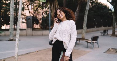 Modern şehir parkında yürürken ve sohbetin tadını çıkarırken kıvırcık saçlı neşeli siyahi kadın gülümsüyor ve arkadaşıyla konuşuyor.