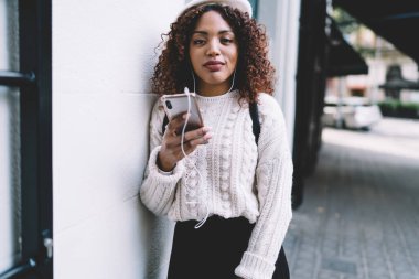 Ciddi kıvırcık saçlı, elinde cep telefonu olan, internetten podcast seçen, akıllı telefon ve kulaklık kullanan güzel Afro-Amerikan kadının portresi.