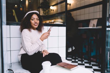 Moda kıyafetli iyimser genç siyahi kadın rahat bir kafede kahve molası verirken cep telefonunda gülümsüyor ve mesajlaşıyor.