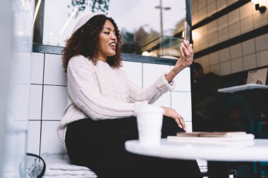 Modern kafede akıllı telefon aracılığıyla görüntülü konuşma yaparken günlük giysiler içinde gülümseyen mutlu Afro-Amerikan kadının alçak açısı.