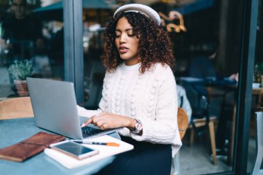 Dizüstü bilgisayarda yazan ciddi koyu tenli kıvırcık kadın web sayfalarına ve bilgilere göz gezdirerek içeriği paylaşıyor. Kafe terasında uzaktan kumandalı iş yapan Afro-Amerikan kadın serbest yazar.