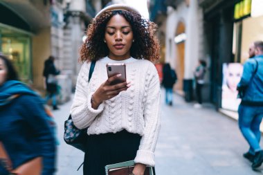 Elinde not defterleri olan beyaz bereli genç bir kadın SMS 'e cevap vermek ve sokak boyunca insanların arasında dolaşmak için telefon kullanıyor.