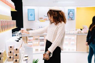 Gündelik giysiler ve bereyle modern güzellik salonunda dikilen ve alışveriş sırasında ürünleri seçen Afro-Amerikan kadınlarının yan görüntüsü