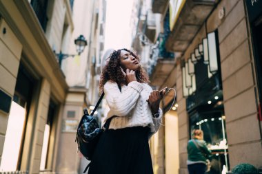 Dar bir sokakta duran ve akıllı telefonuyla kulaklıkla müzik dinleyen, gündelik giysiler ve bereli şık Afro-Amerikan kadınlarının alçak açısı.
