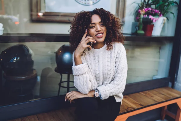 Cheerful dark skinned woman in trendy wear having positive mobile phone conversation using roaming, smiling african american female millennial making smartphone call via international connection