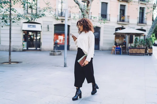 ファッショナブルなアフリカ系アメリカ人女性でホワイトセーター黒パンツとヒール足首ブーツで路上で歩くと日記とメッセージングモバイル — ストック写真