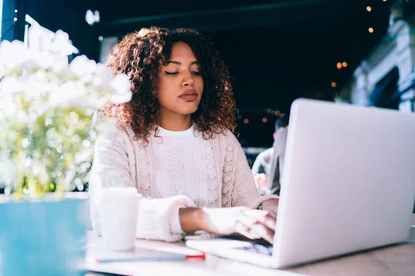 穿着休闲装 心事重重的非洲裔美国女性坐在现代咖啡馆的木桌旁 一边浏览笔记本电脑 一边远程工作 — 图库照片