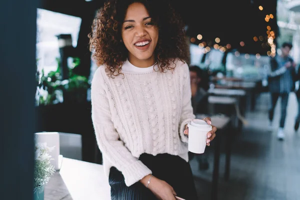 愉快地微笑着 年轻的非洲裔美国女人在辛苦的工作后 一边在现代咖啡馆休息 一边看着相机 一边喝着热外卖咖啡 — 图库照片