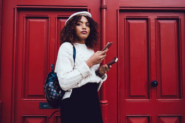 穿着头饰 背着背包 拿着笔记本浏览手机 站在门外的年轻而时髦的非洲裔美国女性 — 图库照片