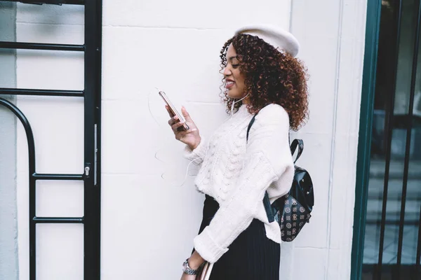 身穿休闲装 头戴贝雷帽 背着背包站在街上浏览智能手机的非洲裔美国女性的侧面照片 — 图库照片