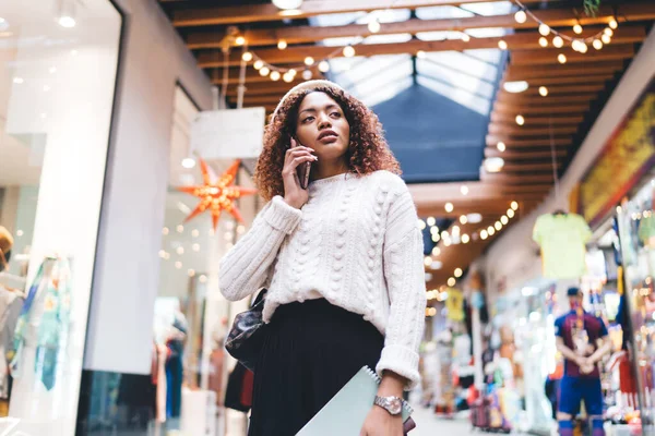 From below of serious African American female in casual clothes and beret walking in modern mall and talking on smartphone during shopping and looking away