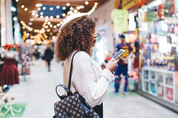 Smiling dark skinned woman in trendy wear walking in mall and checking notification messages on smartphone, happy hipster girl enjoying good news about sales holding mobile phone on shopping