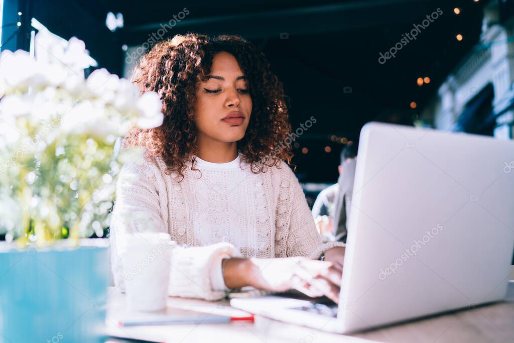Pensive African American female in casual clothes sitting at wooden table in modern cafe while browsing laptop and working remotely
