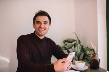 Gündüzleri modern kahve dükkanında kahve içerken kameraya gülümseyen ve akıllı telefon tutan sıradan bir adam.