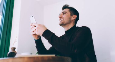 Neşeli Kafkasyalı erkek blogcu elinde dijital akıllı telefonla sevinçle gülümserken cep telefonlu neşeli milenyum esmeriyle kablosuz ağ kurmanın keyfini çıkarıyor.