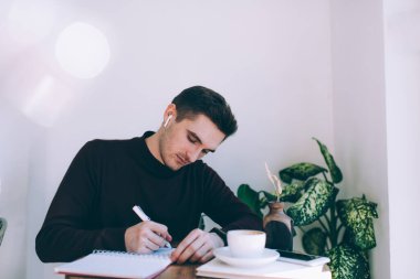 Elektronik kulaklıklı genç beyaz öğrenci kafeterya masasında oturuyor ve üniversite projesi için fikirler yazıyor, kulaklıklarıyla kulaklık takıyor ve içeride öğrenirken ses bilgilerini dinliyor.