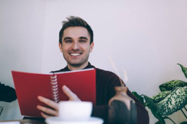 Öğrenme ve öğrenme için eğitim kitabı tutan neşeli beyaz öğrencinin yarım boy portresi, kolej ödevlerini yaparken kameraya gülümseyen planlayıcı not defteriyle neşeli bir erkek.