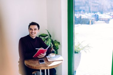 Gündüz kıyafetleri içinde gülümseyen genç adam güneşli bir günde geniş panoramik pencereli yaratıcı çalışma alanında kahve molası üstüne program yapıyor.