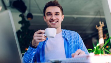 Kafkasyalı neşeli bir adamın yarım uzunluğu kahve fincanı tutuyor ve uzaktan çekim sırasında kameraya gülümsüyor, mutlu erkek milenyumu portresi kafein içeceği ile serbest yaşam tarzının keyfini çıkarıyor.