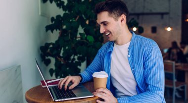 Modern dizüstü bilgisayardaki eğlenceli erkek bilgi işlem programı metni kahve dükkanında mesafelere eğleniyor, paket bardaklı neşeli bir adam uygulama platformu üzerinden online web webinar izliyor.
