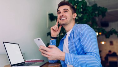 Neşe dolu uzaktan kumandalı erkek çalışanın cep telefonu ve boş ekranlı açık netbook 'u güneş ışığı altında ahşap masaya yaslanmış ve başka yöne bakıyor.