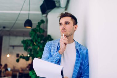 Günümüz ofisinde dikilirken beyaz duvara yaslanan dosyalar ve kağıtlarla sıradan giyinmiş düşünceli bir erkek tarafından.