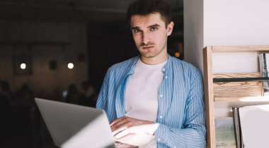 Gündüz vakti elinde dizüstü bilgisayar tutan ve kameraya bakarken beyaz duvara yaslanan ciddi bir adam.