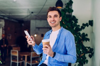 Kameraya gülümseyen ve modern kafeteryada otururken akıllı telefonu elinde tutan, günlük kıyafetlerle, sakallı ve koyu renk saçlı neşeli bir adam.