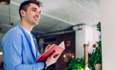 Kafedeki bitkilerin arka planında not defterine not düşerken, siyah saçlı, sıradan elbiseli, dik duran ve başka yöne bakan olumlu genç adam.