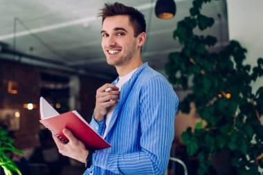 Modern ofiste, yeşil bitkilerin yanında çalışırken kameraya gülümseyen ve kitap okuyan gündelik kıyafetli genç bir erkek.