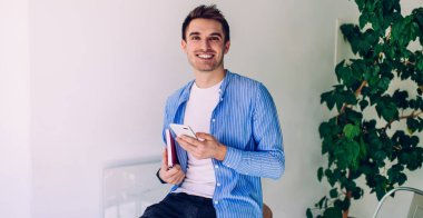 Gündelik kıyafetlerle gezen genç bir adam beyaz modern apartmandaki fabrikanın yanındaki sandalyede otururken elinde not defteri ve akıllı telefon kullanıyor.