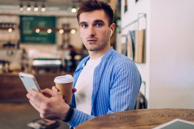 Elektronik kulaklıkla ses şarkılarını seçerken kameraya bakan yakışıklı bir erkeğin portresi. Mobil uygulama kullanarak radyo istasyonunda müzik dinlemek için kahve içen yakışıklı hippi adam.