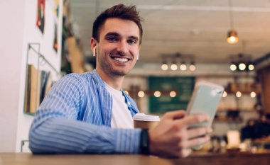 Bluetooth kulaklıklı, 20 yaşında, müzik dinlemek için cep telefonu kullanan neşeli, beyaz bir adamın yarım boy portresi.