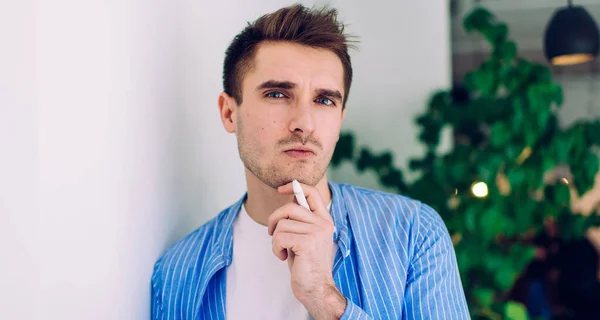 Half length portrait of thoughtful male student with white pen in hand looking at camera while thinking about education idea, contemplative Caucasian man 20s feeling doubt and cogitate indoors