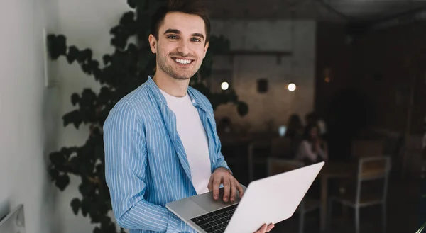 Positive young student in casual wear smiling and looking at camera while standing with laptop and using touchpad in cafe on blurred background