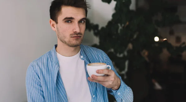Calm young male worker in casual outfit looking at camera while sitting with laptop and drinking coffee in comfortable workplace