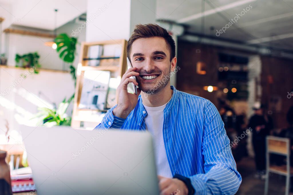 Portrait of funny male IT professional enjoying freelance lifestyle smiling at camera while receiving friendly cellular call, cheerful man making mobile communication for discuss web project