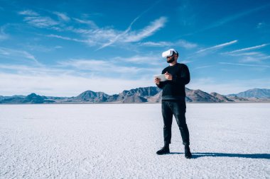 Utah ABD 'nin tuz düzlüklerinde tatil yaparken modern VR gözlükleri ve taşınabilir tablet kullanan gündelik giysiler içinde tam boy erkek gezgin.