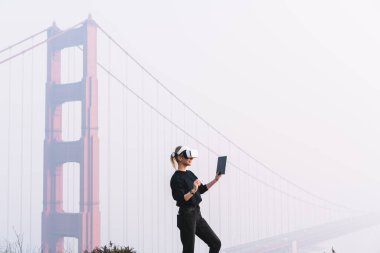 San Francisco Golden Gate Köprüsü yakınlarındaki aletleri test ederken kısa saçlı, sanal gerçeklik gözlüklü kadın tabletteki verileri kontrol ediyor.