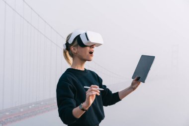 Sisli bir günde San Francisco 'da Golden Gate yakınlarında dururken sanal gerçeklik deneyimi deneyimi yaşayan, VR kulaklık takan, dijital kalem ve tablet kullanan inanılmaz bir kadın.