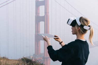 San Francisco 'da Golden Gate' in yanında elinde tablet ve stille dikilirken günlük kazaklı sarışın bir kadının VR gözlük kullanışının yan görüntüsü.