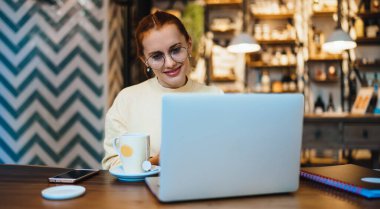 Klasik gözlüklü mutlu bayan öğrenci, kahve dükkanında boş zamanlarında halka açık internet bağlantısını kullanarak milenyum boyunca web sitesinde pozitif video izliyor. Teknoloji kavramı.