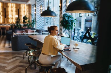 Barcelona 'da modern bir kafede elinde bir kupa çay, mobil ve dizüstü bilgisayarla uzun tahta masada oturan, günlük kıyafetli, gözlüklü, gülümseyen genç bir kadının yan görüntüsü.