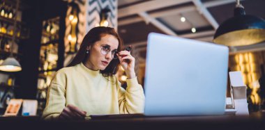 Kablosuz internete bağlı dizüstü bilgisayardan öğrenme sırasında çevrimiçi webinar izleyen çekici beyaz kadın, klasik gözlüklü güzel bir kadın ekrana bakarak medya dosyaları indiriyor.