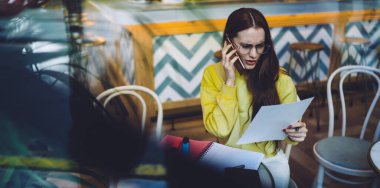 Güzel, genç ve serbest çalışan bir bayan, rahat bir kafede oturmuş, akıllı telefon konuşmaları yaparken, iş belgelerini okuyor ve kaş çatıyor.