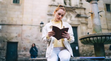 Barcelona City 'deki eski meydanda fıskiyenin yanında otururken not defterinde günlük kıyafetlerle notlar okuyan düşük açılı genç bir kadın.