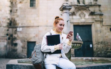 Sırt çantalı ve defterli neşeli genç bayan Barcelona caddesindeki eski bir çeşmede otururken gülümsüyor ve akıllı telefonuna göz atıyor.